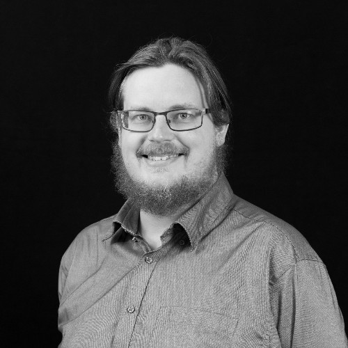 Headshot of a smiling man with a beard and glasses, wearing a smart shirt.